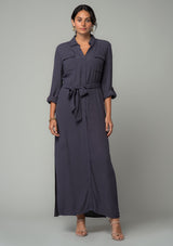 [Color: Charcoal] A front facing image of a brunette model wearing a relaxed fit charcoal grey maxi shirt dress made from a lightweight crepe. With long rolled sleeves, a button tab sleeve closure, two front pockets, side pockets at the hip, and a self tie waist belt. 