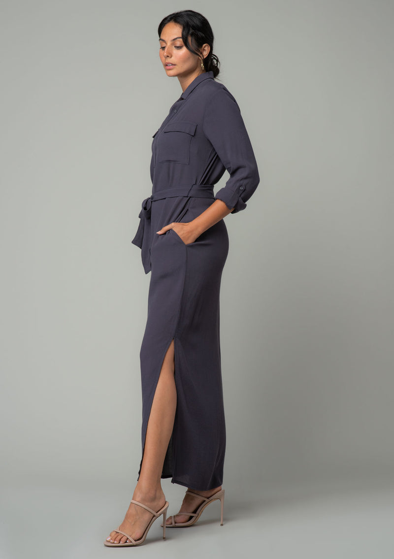 [Color: Charcoal] A side facing image of a brunette model wearing a relaxed fit charcoal grey maxi shirt dress made from a lightweight crepe. With long rolled sleeves, a button tab sleeve closure, two front pockets, side pockets at the hip, and a self tie waist belt. 