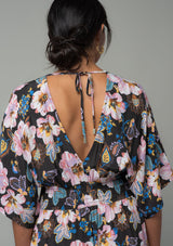[Color: Charcoal/Rose] A close up back facing image of a brunette model wearing a charcoal grey and pink floral print bohemian resort maxi dress with half length kimono sleeves, an open back with tie closure, and a smocked elastic waist. 