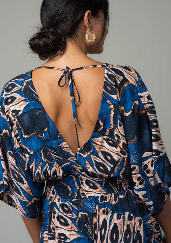 [Color: Cobalt/Tan] A close up back facing image of a brunette model wearing a navy blue and tan butterfly wing printed resort maxi dress with half length kimono sleeves, a smocked elastic waist, an open back with tie closure, and a sexy surplice v neckline. 