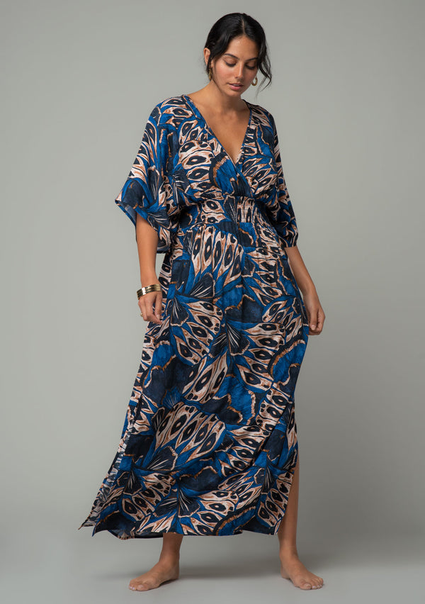 [Color: Cobalt/Tan] A front facing image of a brunette model wearing a navy blue and tan butterfly wing printed resort maxi dress with half length kimono sleeves, a smocked elastic waist, an open back with tie closure, and a sexy surplice v neckline. 