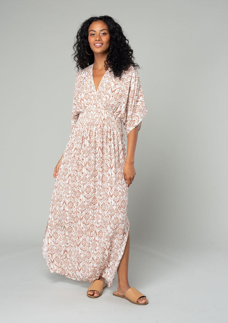 [Color: Ivory/Sand] A front facing image of a brunette model wearing a best selling resort maxi dress in an ivory and brown bohemian diamond print. With short kimono sleeves, a smocked elastic waist, a v neckline, side slits, and an open back with tie closure. 