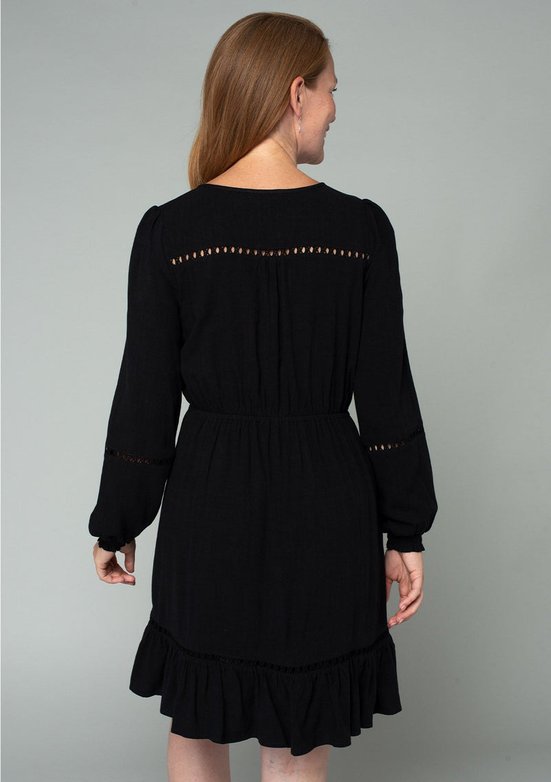 [Color: Black] A back facing image of a red headed model wearing a black linen blend mini dress. A classic bohemian dress with voluminous long sleeves, a self covered loop button front, an elastic waist with tassel tie accent, and crochet trim throughout. 