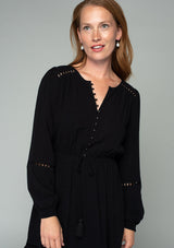 [Color: Black] A close up front facing image of a red headed model wearing a black linen blend mini dress. A classic bohemian dress with voluminous long sleeves, a self covered loop button front, an elastic waist with tassel tie accent, and crochet trim throughout. 