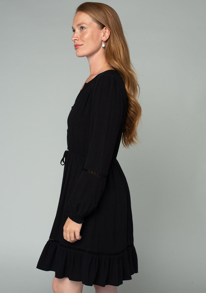 [Color: Black] A side facing image of a red headed model wearing a black linen blend mini dress. A classic bohemian dress with voluminous long sleeves, a self covered loop button front, an elastic waist with tassel tie accent, and crochet trim throughout. 