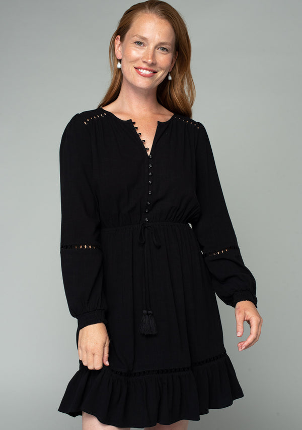 [Color: Black] A front facing image of a red headed model wearing a black linen blend mini dress. A classic bohemian dress with voluminous long sleeves, a self covered loop button front, an elastic waist with tassel tie accent, and crochet trim throughout. 