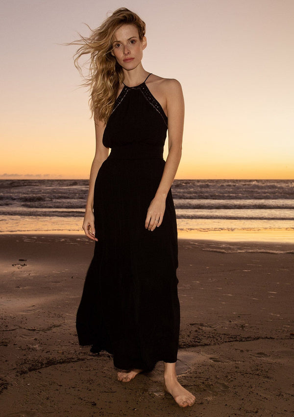 [Color: Black] A front facing image of a blonde model on the beach wearing a bohemian black halter maxi dress. A holiday maxi dress with thin spaghetti straps, a back keyhole, a halter neckline with adjustable back tassel ties, light catching beaded accents, a half smocked elastic waist at the back, and a long flowy skirt.