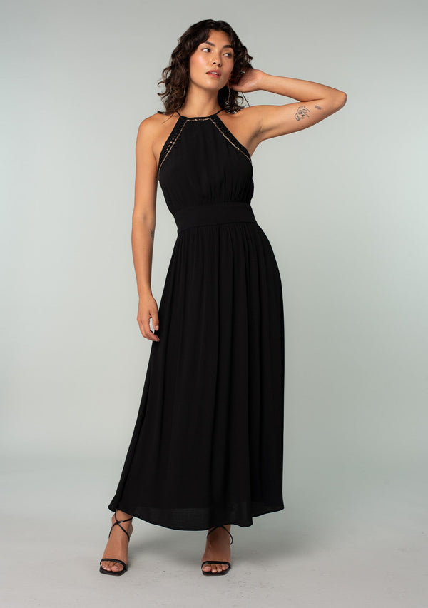 [Color: Black] A front facing image of a brunette model wearing a bohemian black halter maxi dress. A holiday maxi dress with thin spaghetti straps, a back keyhole, a halter neckline with adjustable back tassel ties, light catching beaded accents, a half smocked elastic waist at the back, and a long flowy skirt. 