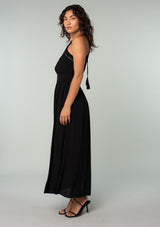 [Color: Black] A side facing image of a brunette model wearing a bohemian black halter maxi dress. A holiday maxi dress with thin spaghetti straps, a back keyhole, a halter neckline with adjustable back tassel ties, light catching beaded accents, a half smocked elastic waist at the back, and a long flowy skirt. 
