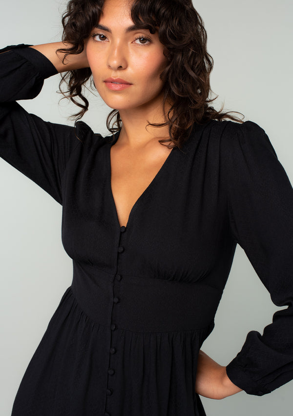 [Color: Black] A close up front facing image of a brunette model wearing a black long sleeve mini dress. Perfect for the holidays, with a self covered button up front, a v neckline, and a half smocked elastic waist at the back.