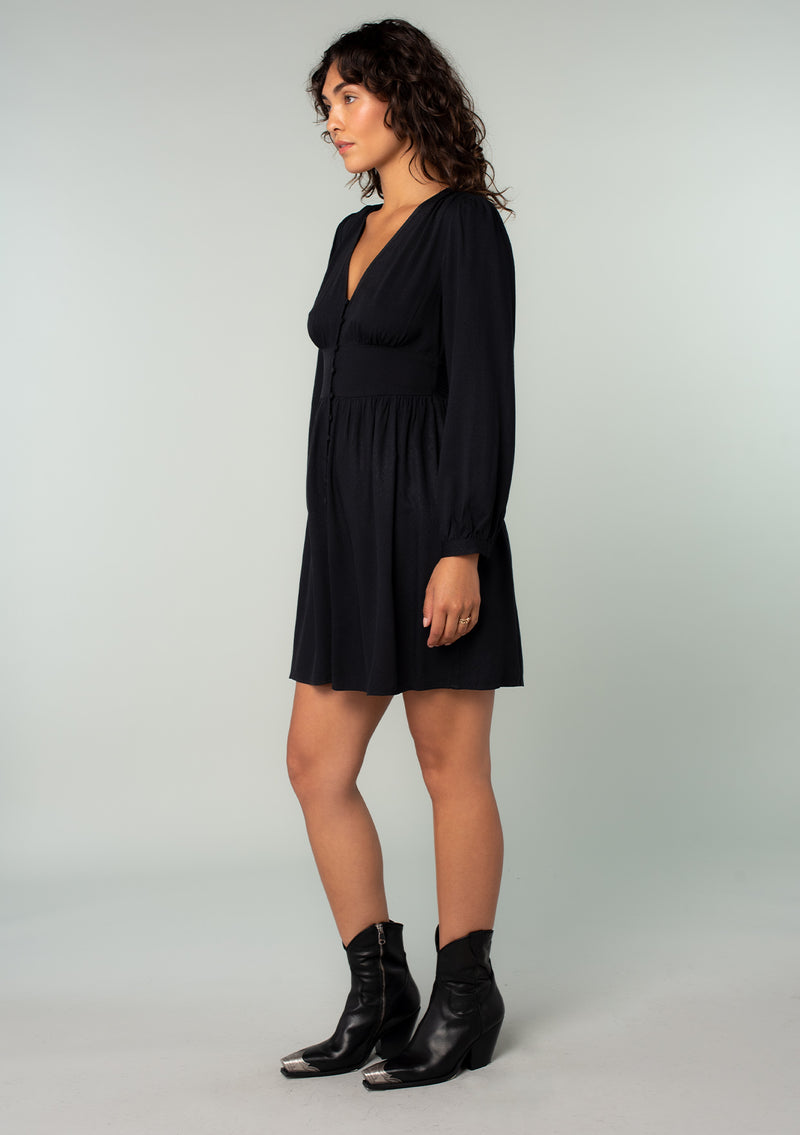 [Color: Black] A side facing image of a brunette model wearing a black long sleeve mini dress. Perfect for the holidays, with a self covered button up front, a v neckline, and a half smocked elastic waist at the back.