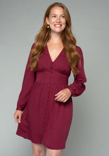 [Color: Wine] A half body front facing image of a red headed model wearing a wine red long sleeve mini dress. Perfect for the holidays, with a self covered button up front, a v neckline, and a half smocked elastic waist at the back. 