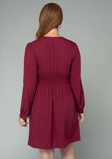 [Color: Wine] A back facing image of a red headed model wearing a wine red long sleeve mini dress. Perfect for the holidays, with a self covered button up front, a v neckline, and a half smocked elastic waist at the back. 