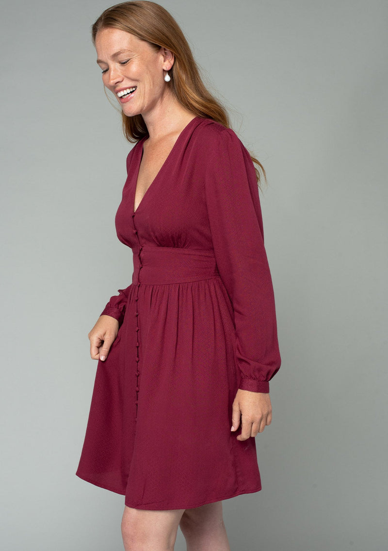 [Color: Wine] A side facing image of a red headed model wearing a wine red long sleeve mini dress. Perfect for the holidays, with a self covered button up front, a v neckline, and a half smocked elastic waist at the back. 