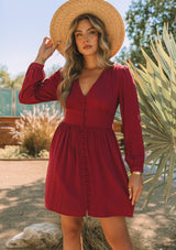 [Color: Wine] A front facing image of a blonde model wearing a wine red long sleeve mini dress. Perfect for the holidays, with a self covered button up front, a v neckline, and a half smocked elastic waist at the back.