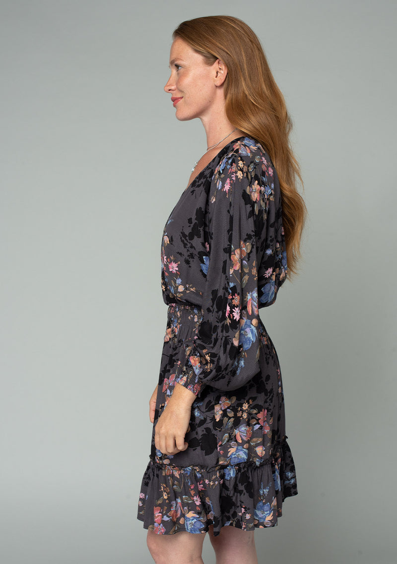 [Color: Grey/Dusty Blue] A side facing image of a red headed model wearing a bohemian fall mini dress in a grey and blue floral print. With voluminous long sleeves, a flowy ruffle trimmed tiered skirt, a smocked elastic waist, and a back keyhole with a tassel tie closure. 