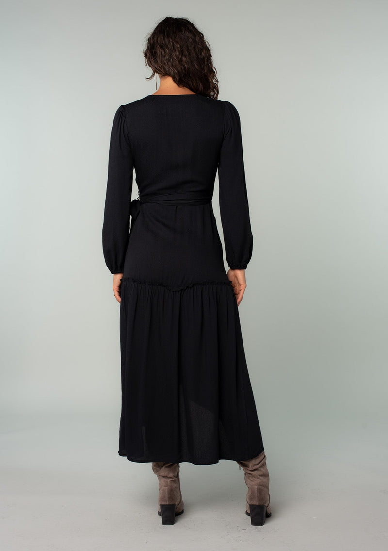 [Color: Black] A back facing image of a brunette model wearing a black maxi wrap dress with long sleeves, a ruffle trimmed tiered high low hemline, and a side tie closure. 