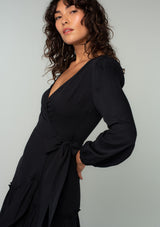 [Color: Black] A close up side facing image of a brunette model wearing a black maxi wrap dress with long sleeves, a ruffle trimmed tiered high low hemline, and a side tie closure. 