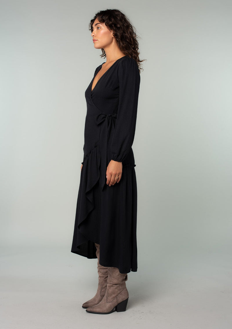 [Color: Black] A side facing image of a brunette model wearing a black maxi wrap dress with long sleeves, a ruffle trimmed tiered high low hemline, and a side tie closure. 