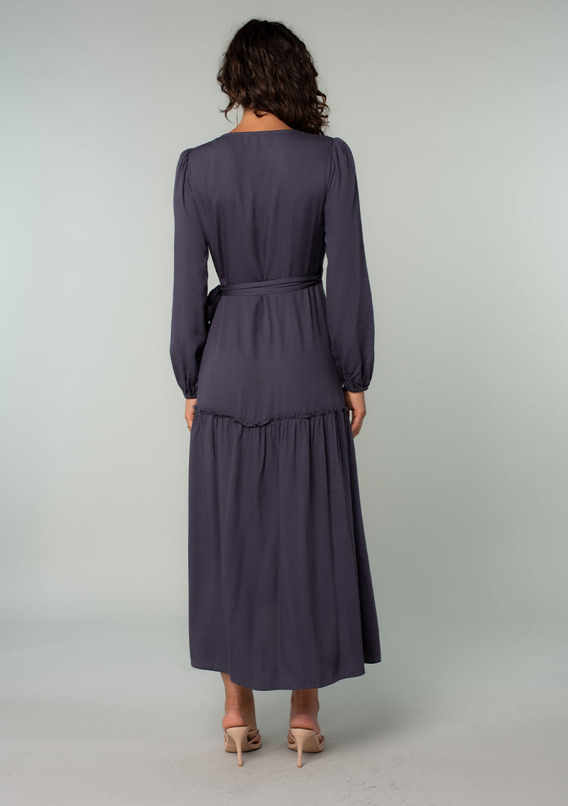 [Color: Periscope Grey] A back facing image of a brunette model wearing a dark grey maxi wrap dress with long sleeves, a ruffle trimmed tiered high low hemline, and a side tie closure. 
