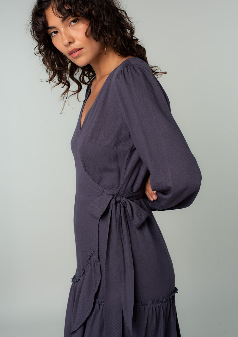 [Color: Periscope Grey] A close up side facing image of a brunette model wearing a dark grey maxi wrap dress with long sleeves, a ruffle trimmed tiered high low hemline, and a side tie closure. 