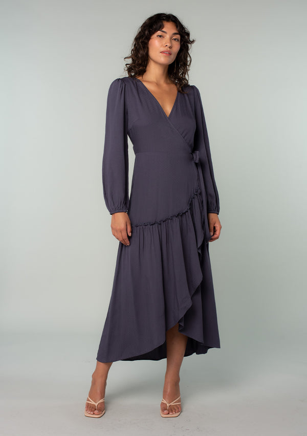 [Color: Periscope Grey] A front facing image of a brunette model wearing a dark grey maxi wrap dress with long sleeves, a ruffle trimmed tiered high low hemline, and a side tie closure. 