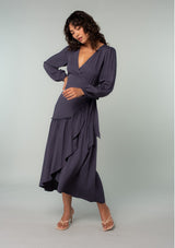 [Color: Periscope Grey] A full body front facing image of a brunette model wearing a dark grey maxi wrap dress with long sleeves, a ruffle trimmed tiered high low hemline, and a side tie closure. 