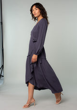 [Color: Periscope Grey] A side facing image of a brunette model wearing a dark grey maxi wrap dress with long sleeves, a ruffle trimmed tiered high low hemline, and a side tie closure. 