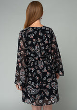 [Color: Black/Dusty Rose] A back facing image of a red headed model wearing a sheer chiffon mini dress in a black and rose pink floral print. With dramatic flared long sleeves, a v neckline, and an elastic waist with a tie accent. 