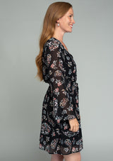 [Color: Black/Dusty Rose] A side facing image of a red headed model wearing a sheer chiffon mini dress in a black and rose pink floral print. With dramatic flared long sleeves, a v neckline, and an elastic waist with a tie accent. 