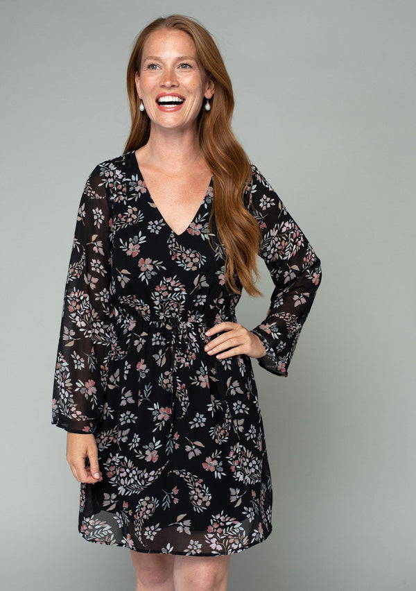 [Color: Black/Dusty Rose] A front facing image of a red headed model wearing a sheer chiffon mini dress in a black and rose pink floral print. With dramatic flared long sleeves, a v neckline, and an elastic waist with a tie accent. 