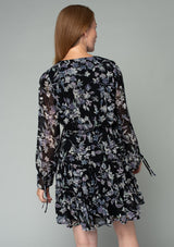 [Color: Black/Lavender] A back facing image of a red headed model wearing a sheer chiffon bohemian mini dress in a black and lavender purple floral print. With a ruffle trimmed tiered skirt, an elastic waist, a v neckline, and long flared sleeves with adjustable drawstring wrist ties. 