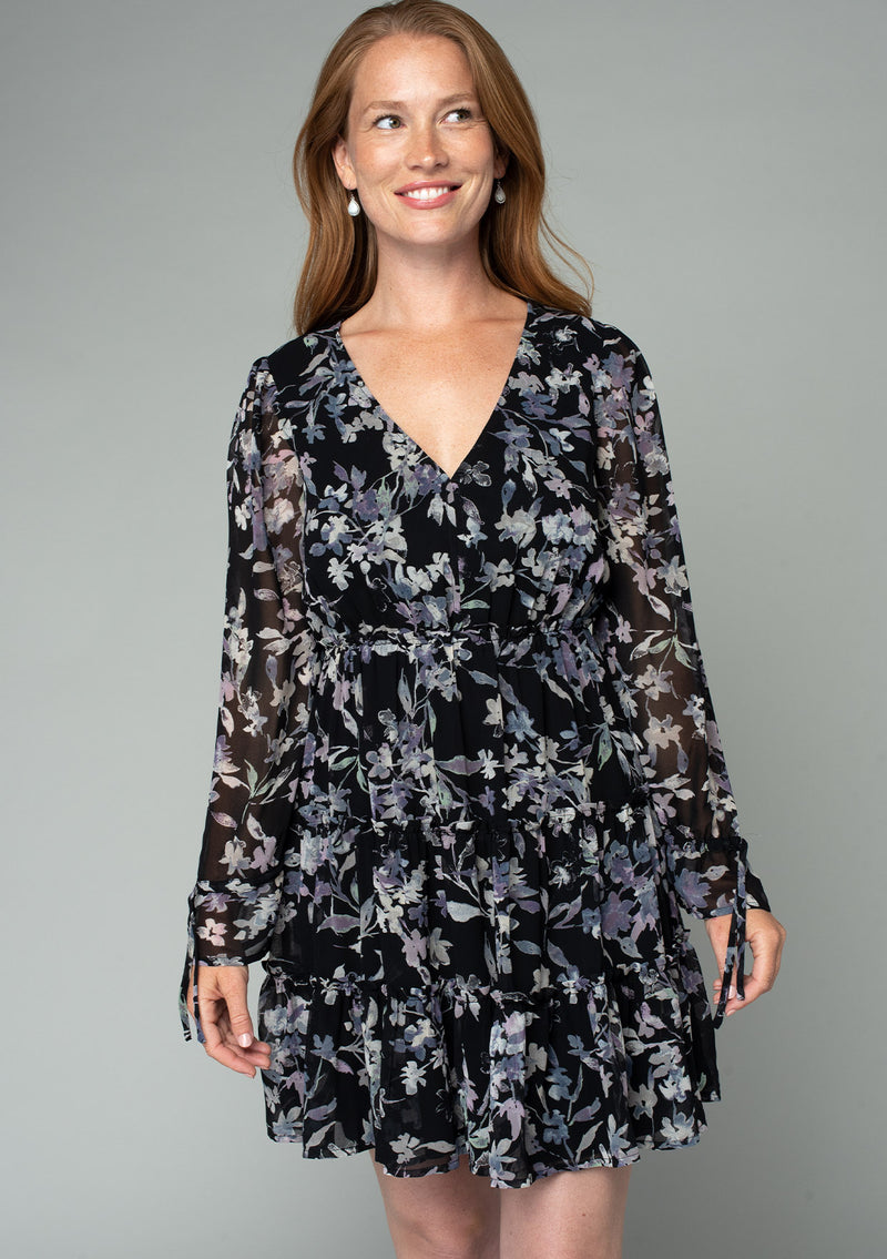 [Color: Black/Lavender] A front facing image of a red headed model wearing a sheer chiffon bohemian mini dress in a black and lavender purple floral print. With a ruffle trimmed tiered skirt, an elastic waist, a v neckline, and long flared sleeves with adjustable drawstring wrist ties. 