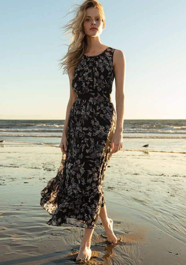 [Color: Black/Lavender] A front facing image of a blonde model on the beach wearing a black and lavender purple floral print chiffon maxi dress. A sleeveless holiday maxi dress with a long tiered skirt and an open back keyhole with tie closure.