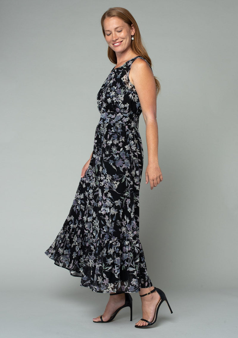 [Color: Black/Lavender] A side facing image of a red headed model wearing a black and lavender purple floral print chiffon maxi dress. A sleeveless holiday maxi dress with a long tiered skirt and an open back keyhole with tie closure. 