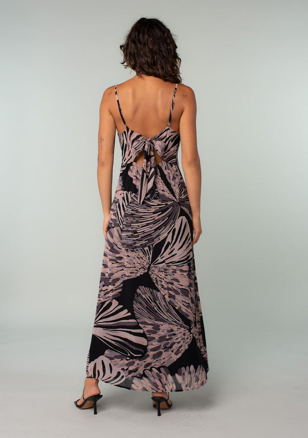 [Color: Black/Natural] A back facing image of a brunette model wearing a chiffon maxi slip dress in a black and natural abstract butterfly wing print. With adjustable spaghetti straps, a v neckline, a front slit, and an adjustable tie back detail. 