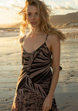 [Color: Black/Natural] A front facing image of a blonde model on the beach wearing a chiffon maxi slip dress in a black and natural abstract butterfly wing print. With adjustable spaghetti straps, a v neckline, a front slit, and an adjustable tie back detail.