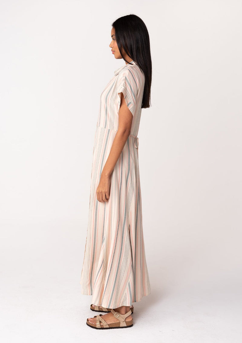 [Color: Natural/Sage] A side facing image of a brunette model wearing a bohemian flowy maxi shirt dress in a natural and sage green stripe. With short cuffed sleeves, a button front, a collared neckline, side pockets, a paneled skirt, and a back waist tie.