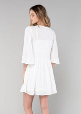 [Color: Vanilla] A back facing image of a blonde model wearing a bohemian white flowy baby doll mini dress. A spring dress with half length flared sleeves, a v neckline, a self covered button front, and lattice trim. 