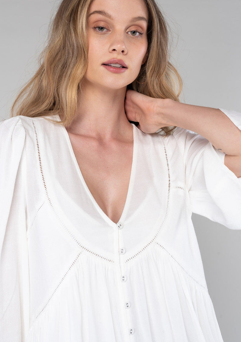 [Color: Vanilla] A close up front facing image of a blonde model wearing a bohemian white flowy baby doll mini dress. A spring dress with half length flared sleeves, a v neckline, a self covered button front, and lattice trim. 
