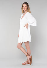 [Color: Vanilla] A side facing image of a blonde model wearing a bohemian white flowy baby doll mini dress. A spring dress with half length flared sleeves, a v neckline, a self covered button front, and lattice trim. 