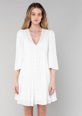 [Color: Vanilla] A front facing image of a blonde model wearing a bohemian white flowy baby doll mini dress. A spring dress with half length flared sleeves, a v neckline, a self covered button front, and lattice trim. 