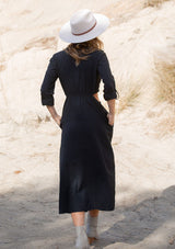 [Color: Black] A back facing image of a blonde model wearing a black cotton mid length shirt dress with long rolled sleeves, a button front, side pockets, and sexy side cutout waist details.