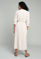 [Color: Stone] A back facing image of a brunette model wearing a natural cotton mid length shirt dress with long rolled sleeves, a button front, side pockets, and sexy side cutout waist details.