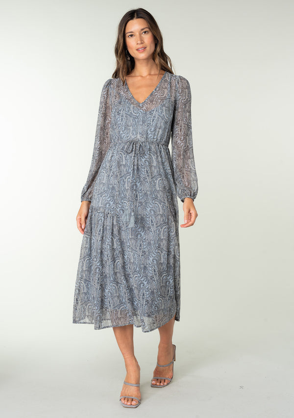 [Color: Grey/Natural] A full body front facing image of a brunette model wearing a sheer chiffon maxi dress in a grey and natural paisley print. With sheer long sleeves, a drawstring waist with tassel ties, a v neckline, and a paneled tiered flowy skirt. 