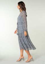 [Color: Grey/Natural] A side facing image of a brunette model wearing a sheer chiffon maxi dress in a grey and natural paisley print. With sheer long sleeves, a drawstring waist with tassel ties, a v neckline, and a paneled tiered flowy skirt. 