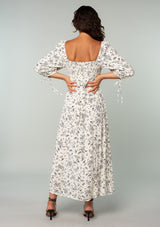 [Color: Natural/Black] A back facing image of a brunette model wearing a bohemian mid length dress in an off white and black ditsy floral print. With three quarter length sleeves, wrist ties, a slim fit smocked bodice, a square neckline, and an empire waist. 