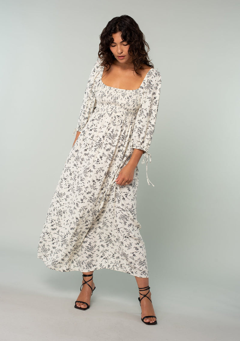 [Color: Natural/Black] A front facing image of a brunette model wearing a bohemian mid length dress in an off white and black ditsy floral print. With three quarter length sleeves, wrist ties, a slim fit smocked bodice, a square neckline, and an empire waist. 