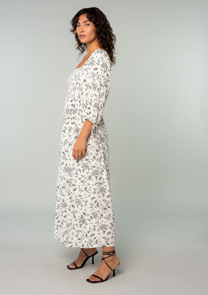 [Color: Natural/Black] A side facing image of a brunette model wearing a bohemian mid length dress in an off white and black ditsy floral print. With three quarter length sleeves, wrist ties, a slim fit smocked bodice, a square neckline, and an empire waist. 