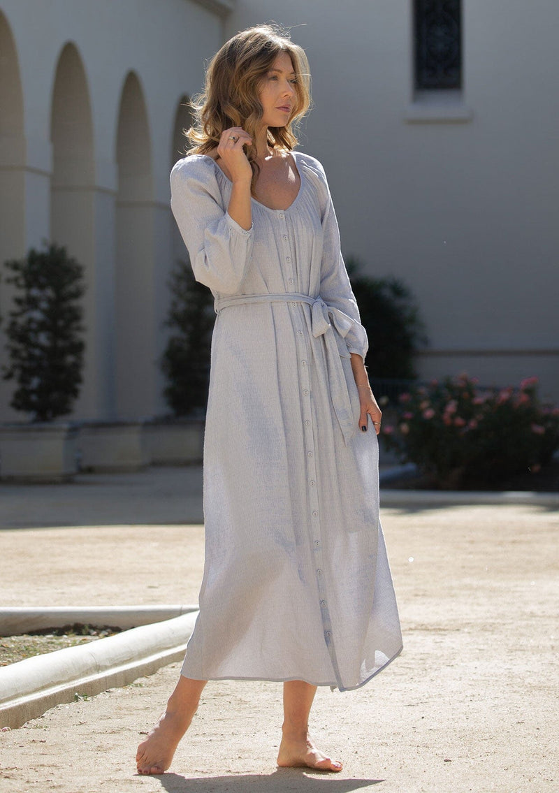 [Color: Dusty Lilac] A front facing image of a blonde model wearing a classic light purple bohemian maxi dress. With long sleeves, a round neckline, a button front, side pockets, and a self tie waist belt.